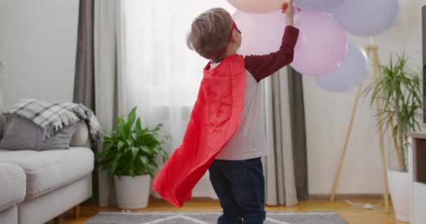 Süper kahraman kostümlü küçük bir çocuk rahat bir oturma odasında renkli balonlarla oynuyor. Yüksek kalite 4k görüntü - Video, Çekim