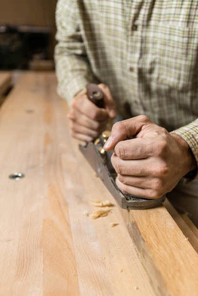 Photo verticale les mains d'un artisan guident habilement un plan à main sur une planche de bois sur un établi, créant des copeaux délicats dans un atelier bien éclairé. Concept d'entreprise. - Photo, image