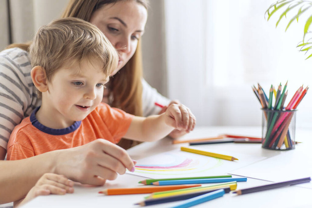 Un niño y su madre comparten un momento de unión mientras dibujan con lápices de colores en casa, con una planta en el fondo. - Foto, imagen