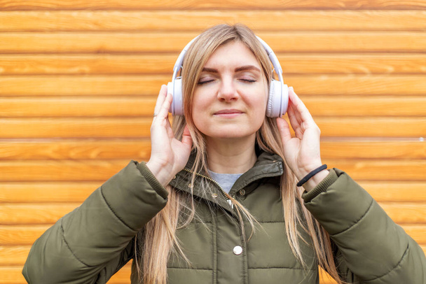 Gözü kapalı genç bir kadın ahşap bir arka planda beyaz kulaklıkla müzik dinliyor.. - Fotoğraf, Görsel