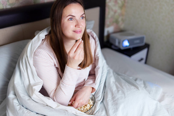 Battaniyeye sarılmış, patlamış mısır yiyip yatakta film izleyen bir kadın. - Fotoğraf, Görsel