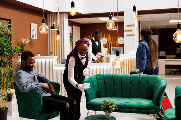 Glimlachende vrouwelijke serveerster serveert cappuccino aan Afrikaans-Amerikaanse hotelgast zet kopje op tafel, brengt vers gebrouwen koffie naar toerist ontspannen in comfortabele lounge in resort - Foto, afbeelding