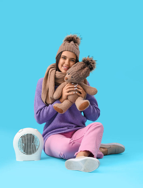 Mulher bonita em roupas de inverno com ursinho de pelúcia e aquecedor de ventilador elétrico no fundo azul - Foto, Imagem