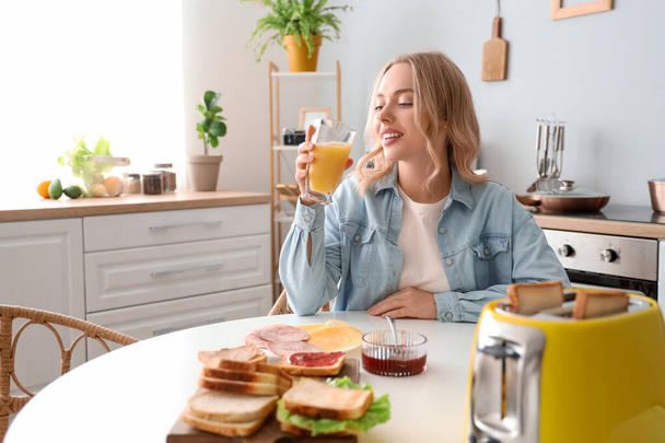 Fiatal, gyönyörű, boldog nő narancslevet iszik finom szendvicsekkel a konyhában. - Fotó, kép