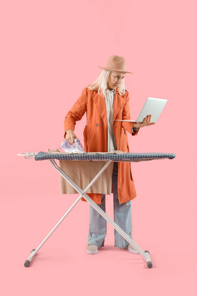 Зріла жінка з ноутбуком для прасування одягу на рожевому фоні. Концепція мультизадачності - Фото, зображення