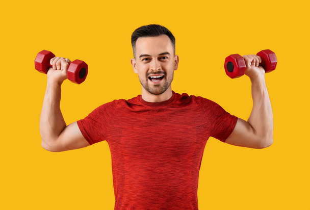 Jóképű fiatal boldog férfi sportruházatban súlyzókkal sárga háttéren - Fotó, kép