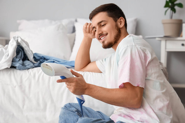 Happy young man with steamer and heap of clean clothes on bed at home - Photo, Image
