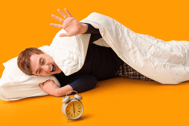 Érzelmes fiatalember takaró és ébresztőóra feküdt narancs háttér - Fotó, kép
