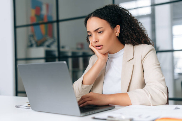 Zdenerwowana brazylijska lub latynoska kręcone włosy kobieta biznesu, dyrektor firmy, dyrektor finansowy, siedzi w nowoczesnym biurze kreatywnym, niestety patrząc na laptopa, studiuje informacje, czyta złe wieści - Zdjęcie, obraz