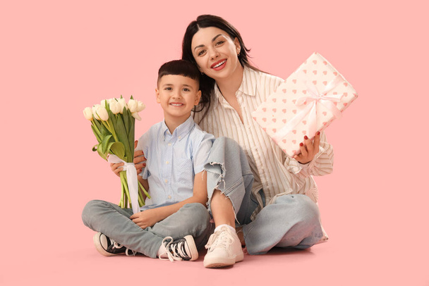 Hermosa mujer con su pequeño hijo sosteniendo tulipanes y regalo sobre fondo rosa. Día Internacional de la Mujer - Foto, imagen