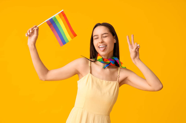 Belle jeune femme avec drapeau LGBT montrant un geste de victoire sur fond jaune - Photo, image