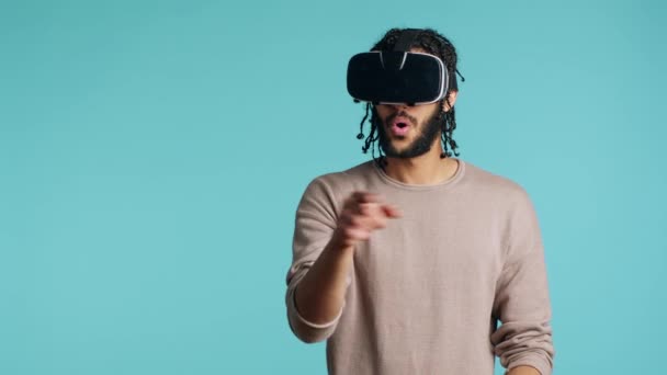 Lähi-idän mies yllään virtuaalitodellisuus kuulokkeet, tekee pyyhkäisemällä eleitä. BIPOC henkilö käyttää korkean teknologian futuristinen moderni VR gadget, eristetty sininen studio tausta, kamera A - Materiaali, video