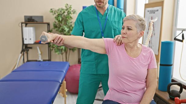 Uma mulher exercita o braço com um halteres em uma clínica de fisioterapia, guiada por um terapeuta do sexo masculino. - Foto, Imagem