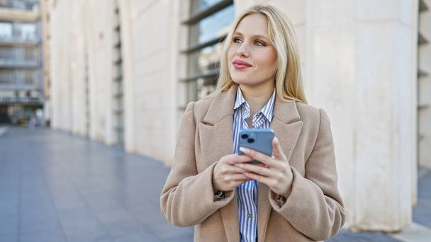 Μια νεαρή γυναίκα που κρατά ένα smartphone στέκεται σε ένα αστικό περιβάλλον, απεικονίζοντας μια αίσθηση της casual συνδεσιμότητας και του σύγχρονου τρόπου ζωής. - Φωτογραφία, εικόνα