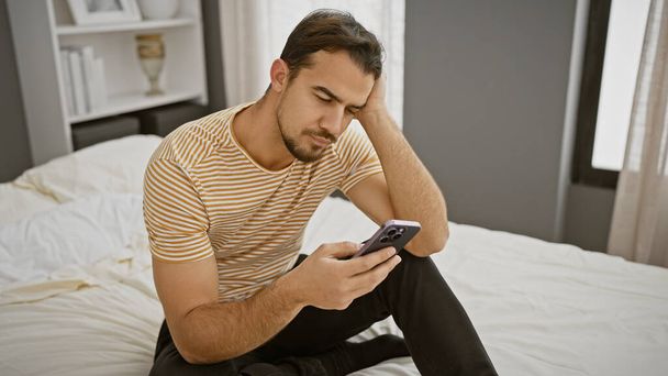 Un bel giovane ispanico con la barba in abiti casual si siede pensieroso sul letto in una camera da letto usando il suo smartphone. - Foto, immagini