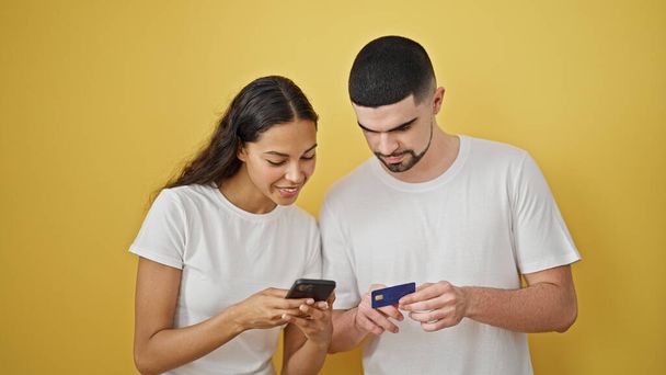 Beautiful couple shopping with smartphone and credit card smiling over isolated yellow background - Photo, Image