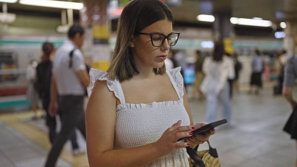 Εκπληκτική Ισπανίδα γυναίκα, με γυαλιά, να στέκεται στον υπόγειο σιδηροδρομικό σταθμό, να περιμένει το ταξίδι της στο μετρό, βυθισμένη στο τηλέφωνό της. - Φωτογραφία, εικόνα
