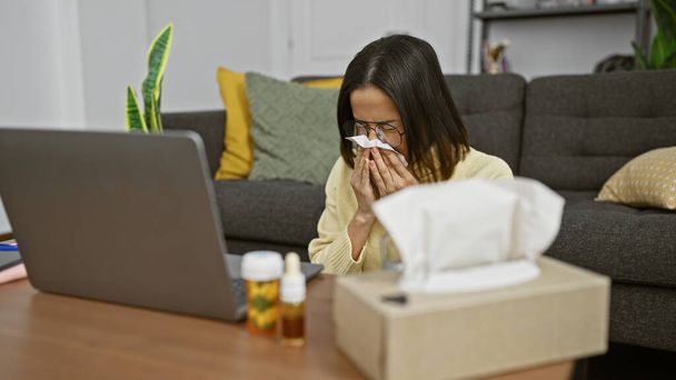 Eine junge hispanische Frau bläst ihre Nase auf ein Gewebe in einem gemütlichen modernen Wohnzimmer, umgeben von Erkältungsmitteln und einem Laptop. - Foto, Bild