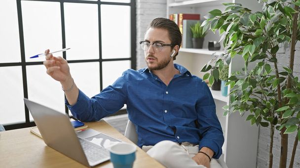Jonge Spaanse man zakenman met behulp van laptop en koptelefoon met pen in het kantoor - Foto, afbeelding