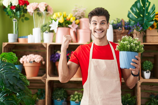 Молодий іспаномовний чоловік, що працює в квітковому магазині, тримає завод з гордістю, святкуючи перемогу і успіх дуже схвильований піднятою рукою  - Фото, зображення