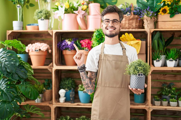 Молодий іспаномовний чоловік з татуюваннями, що працюють у квітковому магазині, тримає рослину усміхненою щасливою, вказуючи рукою і пальцем на бік  - Фото, зображення