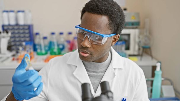 Scienziato afroamericano di sesso maschile che analizza un tubo campione in laboratorio, indossando occhiali di sicurezza e camice da laboratorio. - Foto, immagini