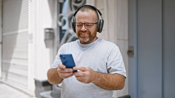 Aantrekkelijke blanke man van middelbare leeftijd, met een serieuze uitdrukking, die gretig een boodschap typt op zijn smartphone terwijl hij buiten op een bankje in de stad zit onder het zonnige zonlicht. - Foto, afbeelding