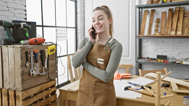 Una giovane donna bionda in un laboratorio di falegnameria che parla al telefono sorridendo in casa. - Foto, immagini