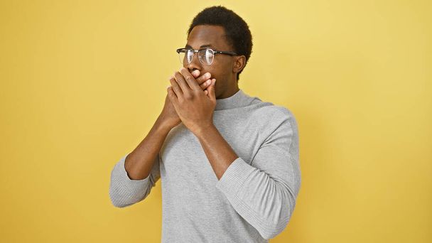 Şaşırmış bir Afro-Amerikan erkeği sarı bir duvar arkasında ağzını kapatıyor, duygularını ve tarzını sergiliyor.. - Fotoğraf, Görsel