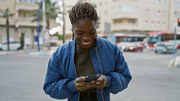 Femme africaine souriante avec des tresses à l'aide d'un smartphone dans une rue de la ville. - Photo, image