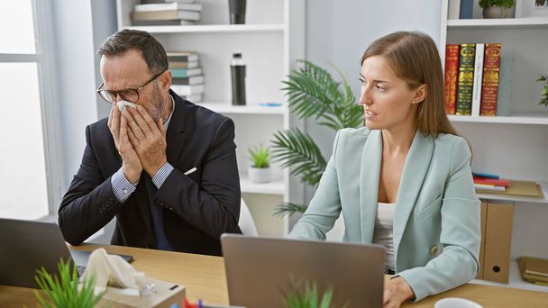 Dva obchodní pracovníci kýchající společně v kanceláři, zatímco pilně pracují na notebooku, jasný výraz onemocnění kancelářské chřipky uvnitř. - Fotografie, Obrázek