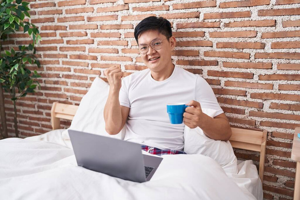 Joven asiático bebiendo café sentado en la cama gritando orgulloso, celebrando la victoria y el éxito muy emocionado con los brazos levantados  - Foto, imagen