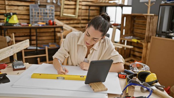 Atractiva joven carpintera hispana absorta en su trabajo, hábilmente dibujando en touchpad mientras está sentada en un acogedor taller de carpintería interior. - Foto, Imagen