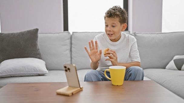 Sevimli sarışın çocuk ciddi ciddi online sohbete daldı, kahvaltı etmek için mobil teknolojiyi kullandı ve oturma odasındaki rahat koltuktan video görüşmesi yaptı. - Fotoğraf, Görsel