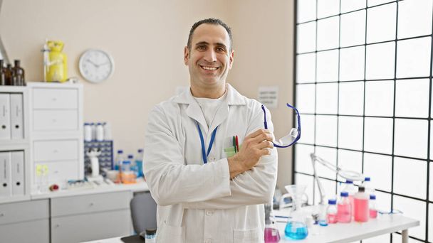 Uomo ispanico sorridente in camice da laboratorio in piedi in una clinica con le braccia incrociate, trasudando fiducia e professionalità. - Foto, immagini