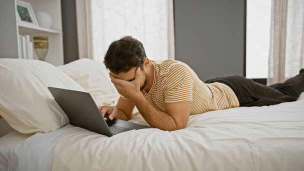 Um homem hispânico estressado deitado em uma cama usando um laptop em um ambiente de quarto moderno, exalando uma sensação de frustração e exaustão. - Foto, Imagem