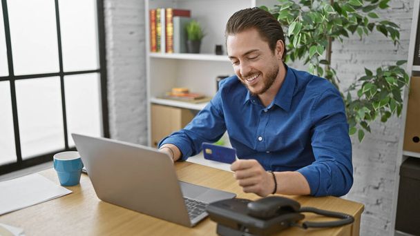 Fiatal spanyol férfi üzletember vásárol laptoppal és hitelkártyával mosolyog az irodában - Fotó, kép
