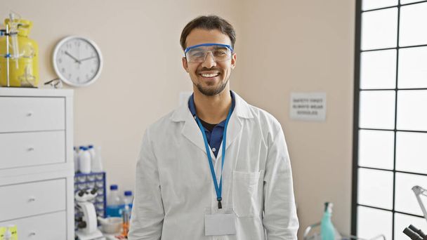 Hymyilevä latino mies yllään lab takki ja turvallisuus suojalasit kirkas laboratorio ympäristössä - Valokuva, kuva
