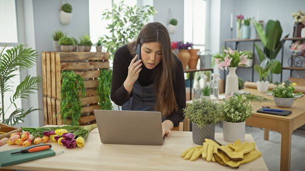 Nuori nainen, joka käyttää kannettavaa tietokonetta ja puhelinta kukkakaupassa, joka on täynnä kasveja, moniajo ja yrittäjyys. - Valokuva, kuva