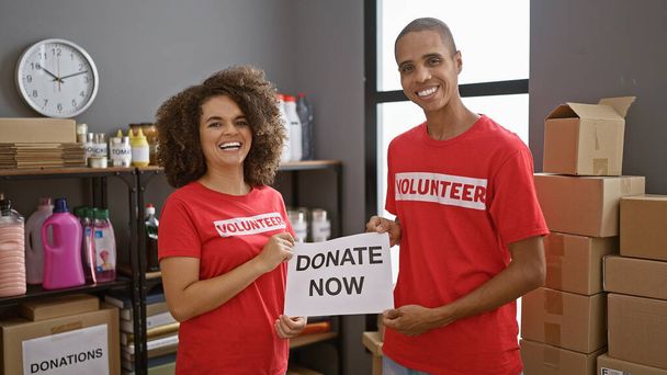Улыбающиеся мужчины и женщины-волонтеры собираются вместе, держа плакат "пожертвуйте сейчас" в центре благотворительного центра, олицетворяя общественный дух и активность - Фото, изображение