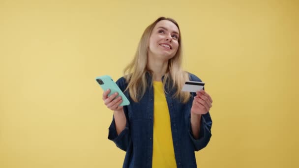 Retrato de una joven rubia feliz mirando la tarjeta de crédito de plástico mientras sostiene el teléfono inteligente aislado sobre el fondo amarillo. Soñadora mujer caucásica reflexiva haciendo la compra y el pago en línea. - Imágenes, Vídeo