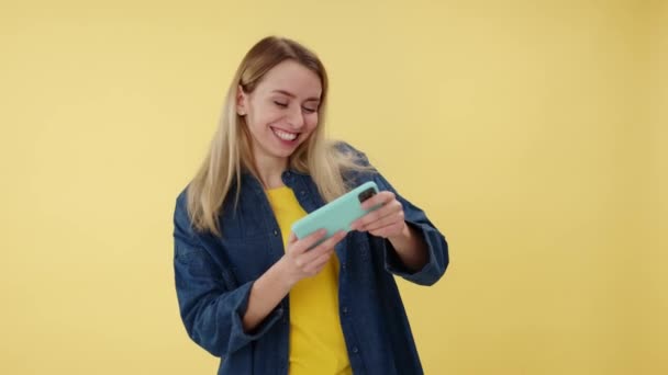 Emotional funny woman enthusiastically playing online video games on modern mobile. Happy cheerful pretty female using smartphone app with drive simulator isolated on yellow studio background. - Footage, Video