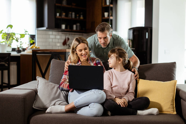 Радісна сім'я з трьох проводить якісний час разом на дивані вітальні, розділяючи мить навколо ноутбука в їх комфортному будинку - Фото, зображення