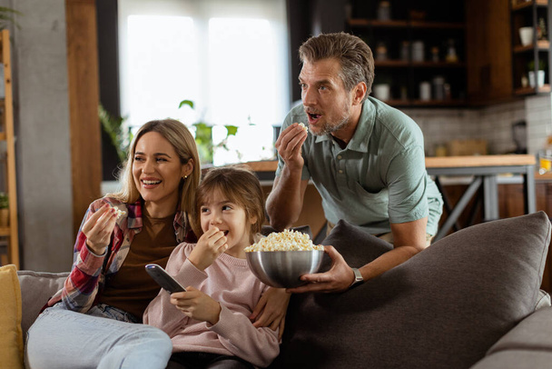 Сім'я з трьох зручно вкладена на диван, їхні обличчя відображають збудження і увагу, коли вони діляться чашею попкорну під час напруженої ночі фільму - Фото, зображення