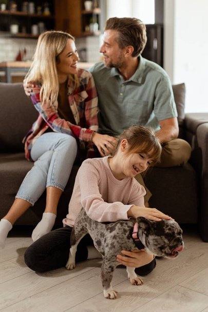 Fröhliche Eltern sehen zu, wie ihre Tochter zu Hause mit einer fröhlichen französischen Bulldogge spielt - Foto, Bild