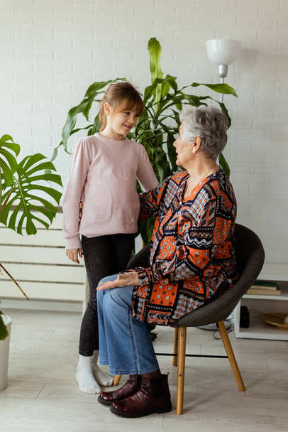 En una habitación suavemente iluminada, una joven se encuentra junto a una planta en maceta, compartiendo un momento de conexión y conversación con su abuela anciana, la alegría y el calor entre ellos palpable - Foto, Imagen