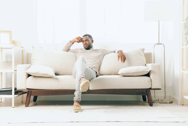 Un homme afro-américain réfléchi assis sur un canapé, réfléchissant sur une vie familiale relaxante et contemplant le bonheur dans un salon moderne - Photo, image