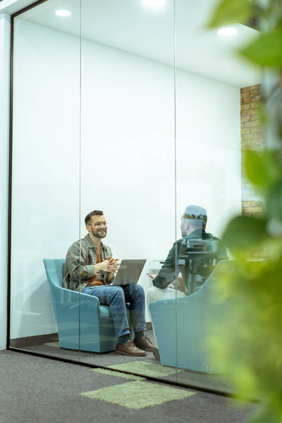Twee professionals voeren een gesprek terwijl ze comfortabel zitten in een kantoorlounge, omgeven door groen en eigentijdse designelementen - Foto, afbeelding