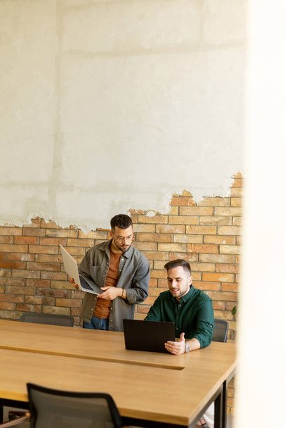 Deux professionnels souriants s'engagent dans une séance de travail collaboratif à une table en bois, leur camaraderie évidente dans un cadre de bureau contemporain avec une toile de fond murale en brique exposée - Photo, image