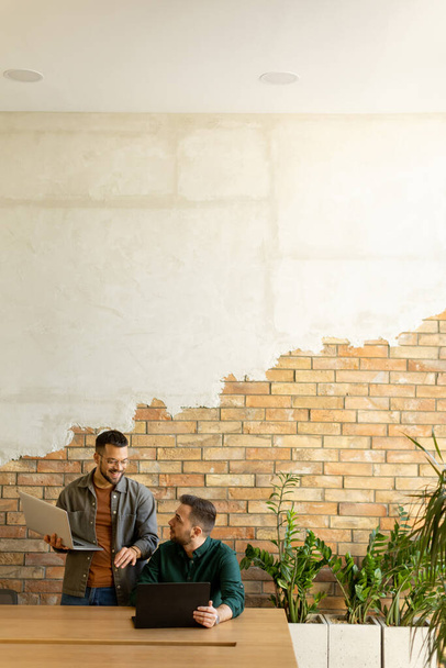 Twee lachende professionals nemen deel aan een gezamenlijke werksessie aan een houten tafel, hun kameraadschap duidelijk in een eigentijdse kantooromgeving met een blootgestelde bakstenen muur achtergrond - Foto, afbeelding